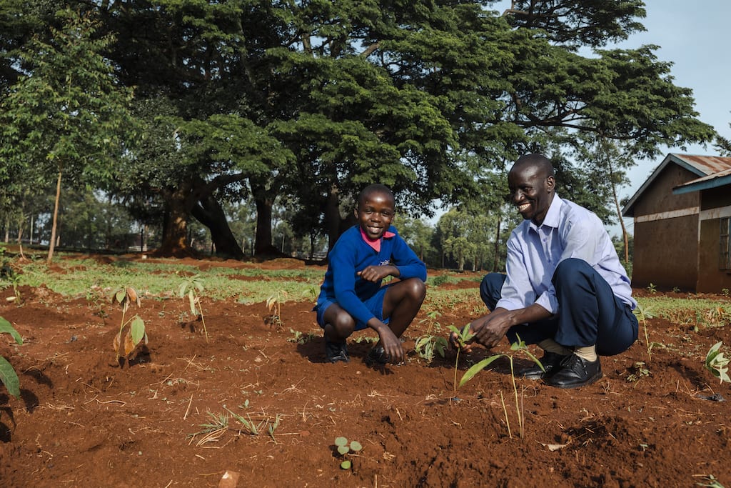 Gilbert, wearing a blue sweater and blue shorts, is with Benson, Center Director, wearing a light blue shirt and dark blue pants. They are kneeling down among newly planted trees.